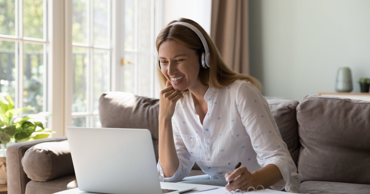 Mulher sentada em um sofá, assistindo a um vídeo em um notebook em cima de uma mesa, tentando aprender inglês sozinha.