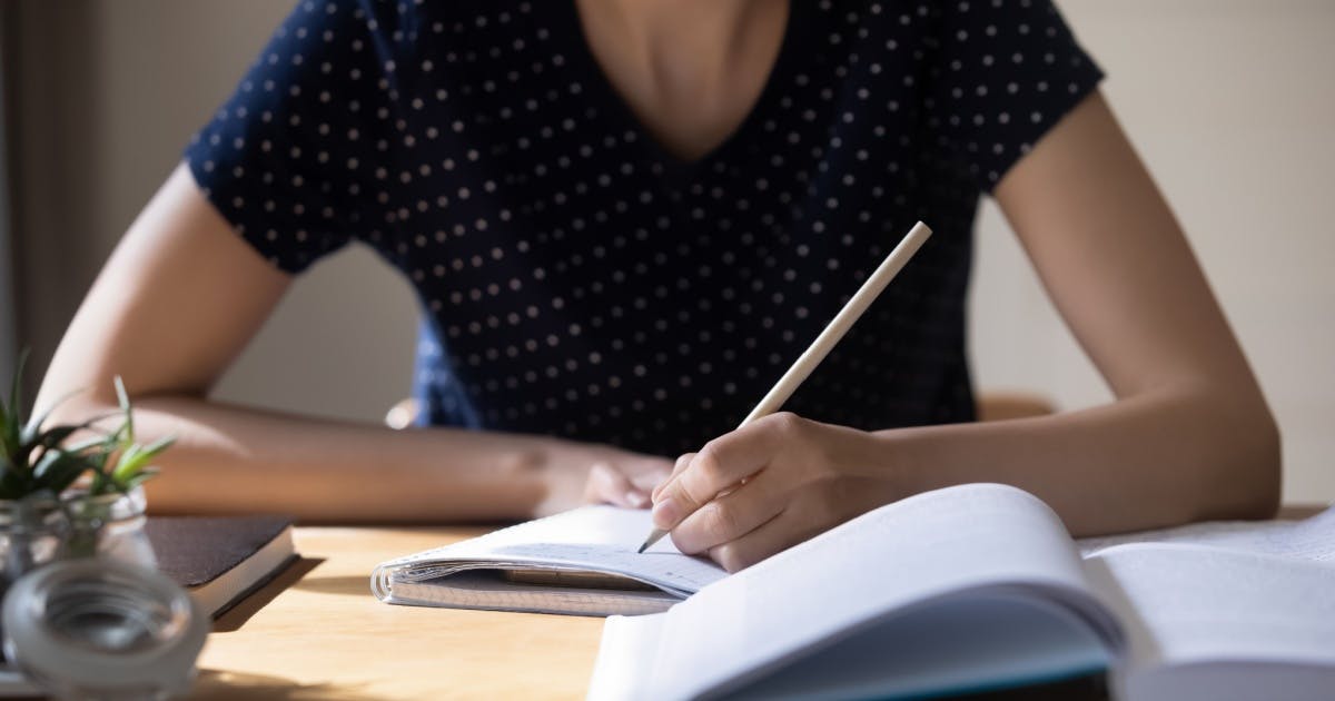 Mulher sentada e apoiada em uma mesa, escrevendo em um caderno com um livro à sua frente, tentando aprender inglês sozinha.