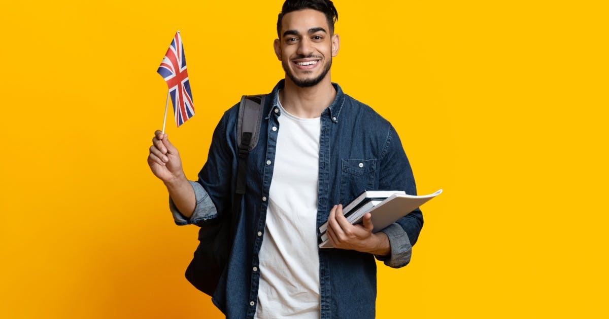 Homem de pé sorrindo, segurando uma pequena bandeira da Grã-Bretanha e alguns livros para tentar aprender inglês sozinho.