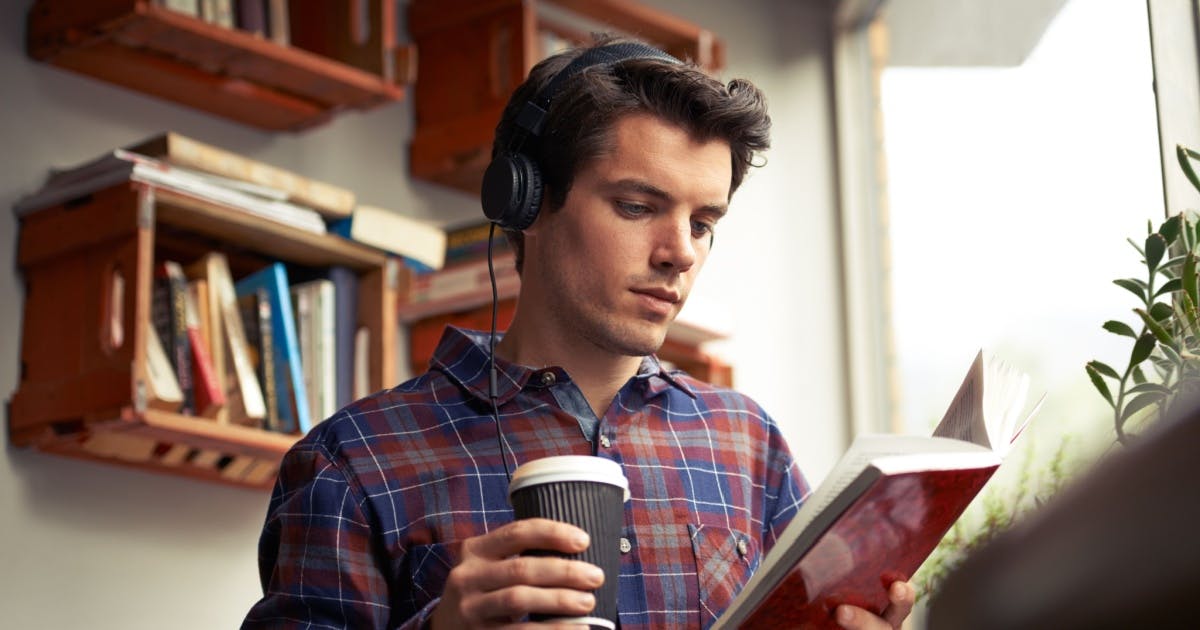 Homem usando um fone de ouvido preto, segurando um copo térmico, enquanto lê um livro ao tentar aprender inglês sozinho.