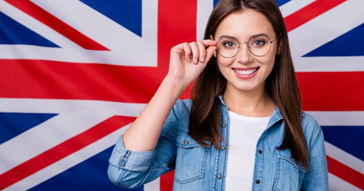 Mulher sorrindo de óculos, usando uma camiseta branca e uma jaqueta jeans, na frente de uma bandeira da Grã-Bretanha.