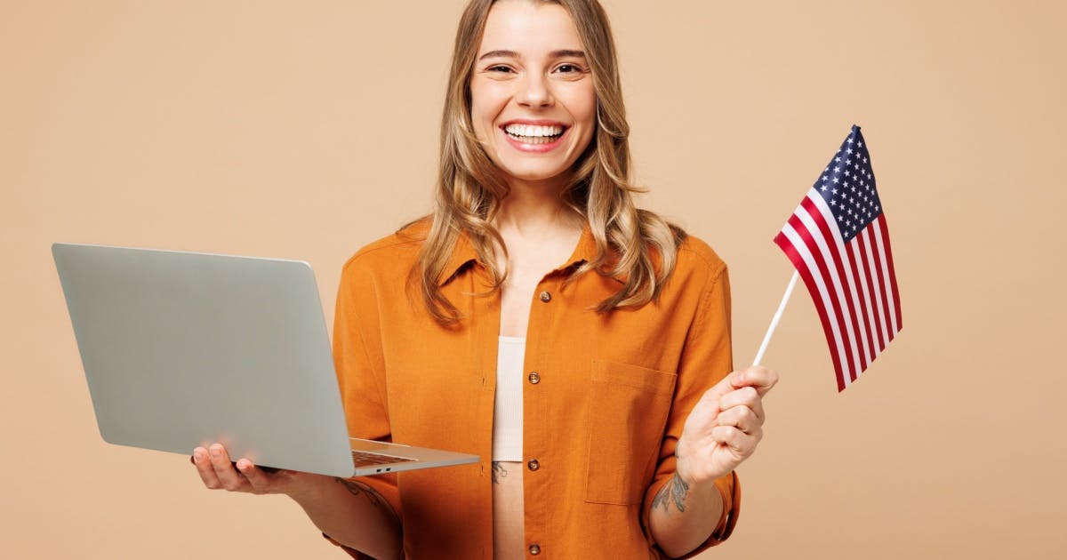 Mulher segurando um pequena bandeira dos Estados Unidos e um notebook aberto, sorrindo por saber como aprender inglês.