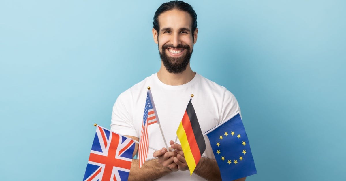 Homem poliglota de camiseta branca, segurando pequenas bandeiras da Grã-Bretanha, Estados Unidos, Alemanha e União Europeia.