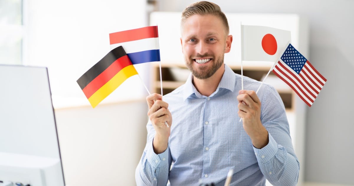 Homem poliglota de camisa social, segurando pequenas bandeiras da Holanda, Estados Unidos, Alemanha e Japão.