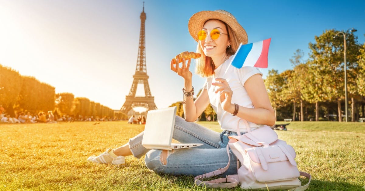 Mulher segurando a bandeira da França, enquanto come um croissant e usa um laptop em Paris, durante aula de um curso de francês.