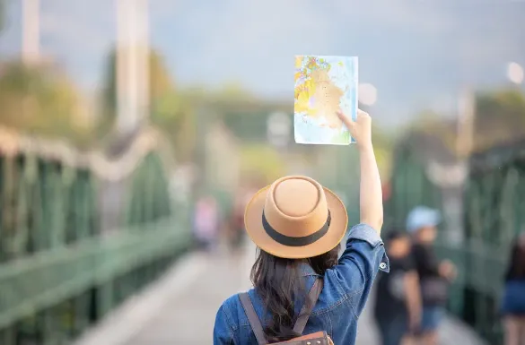 Mulher com chapéu de palha em passeio ao ar livre segura um mapa aberto na mão, com fundo desfocado de pessoas e paisagem urbana
