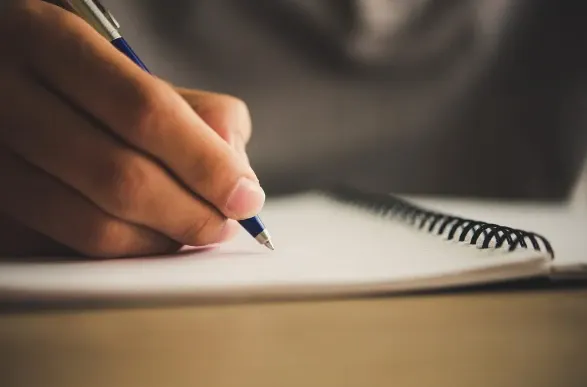 Mão escrevendo com caneta em um caderno aberto sobre mesa, com foco na escrita e ambiente de estudo ou trabalho
