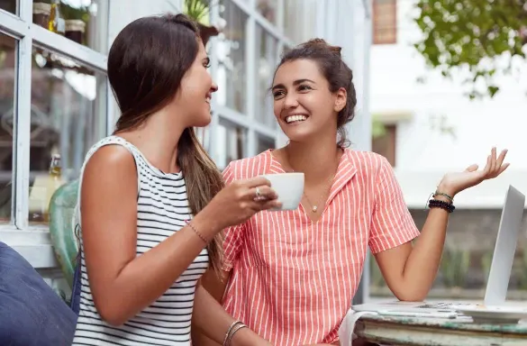 Duas mulheres conversando em uma mesa de café ao ar livre, com uma delas sorrindo enquanto toma uma bebida em xícara branca, em ambiente ensolarado.