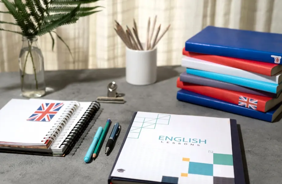 Materiais de estudo de inglês sobre uma mesa: livros com capas azul e vermelha, caderno “ENGLISH LESSONS”, lápis e canetas, além de bandeiras do Reino Unido abertas em um fichário.