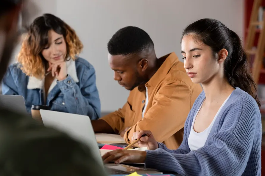 Grupo de pessoas trabalhando e estudando em uma mesa, com uma mulher escrevendo com caneta em folhas coloridas e outros participantes usando computadores em ambiente interno