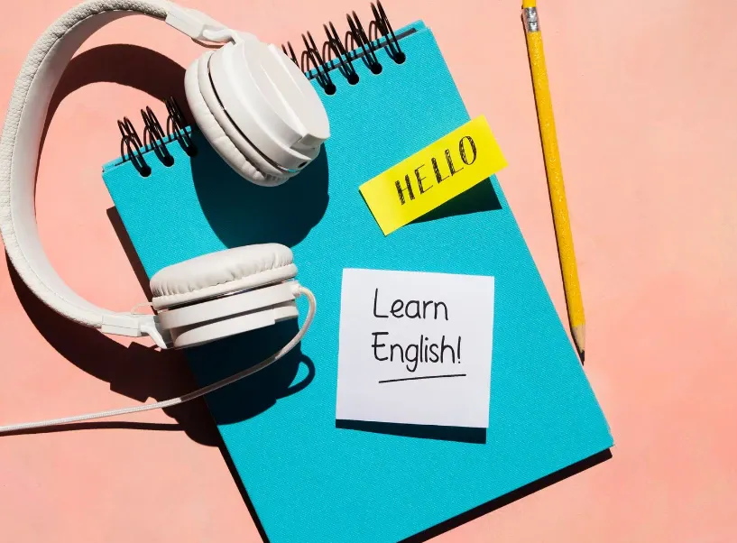 Fones de ouvido brancos sobre um caderno azul com as palavras “HELLO” e um bilhete “Learn English!”, ao lado de um lápis amarelo em fundo rosa.