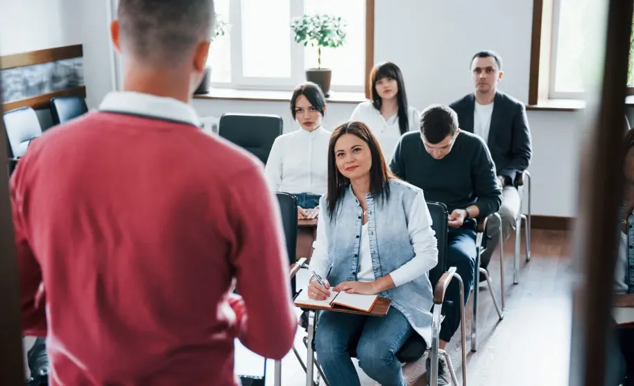 Encontro em sala de reuniões com pessoas sentadas em cadeiras, uma mulher ao centro com caderno e caneta, durante uma conversa ou treinamento profissional em grupo.
