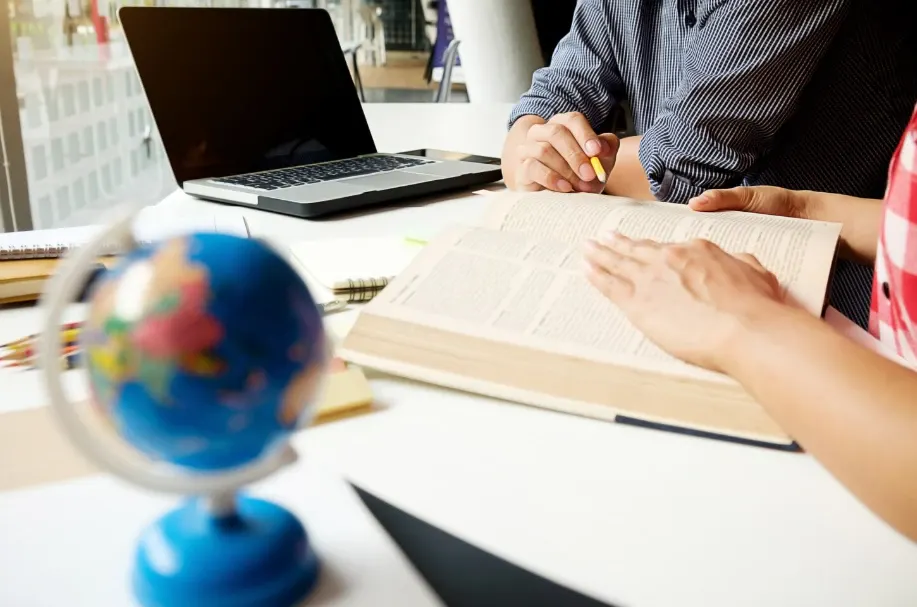 Pessoa estudando e fazendo anotações em um livro aberto sobre a mesa, com notebook ao fundo e um globo desfocado em primeiro plano, em ambiente de estudo.