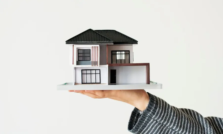 Mão segurando uma maquete de casa com fachada branca e marrom, telhado preto e janelas em destaque, sobre fundo claro, simulando um projeto imobiliário.