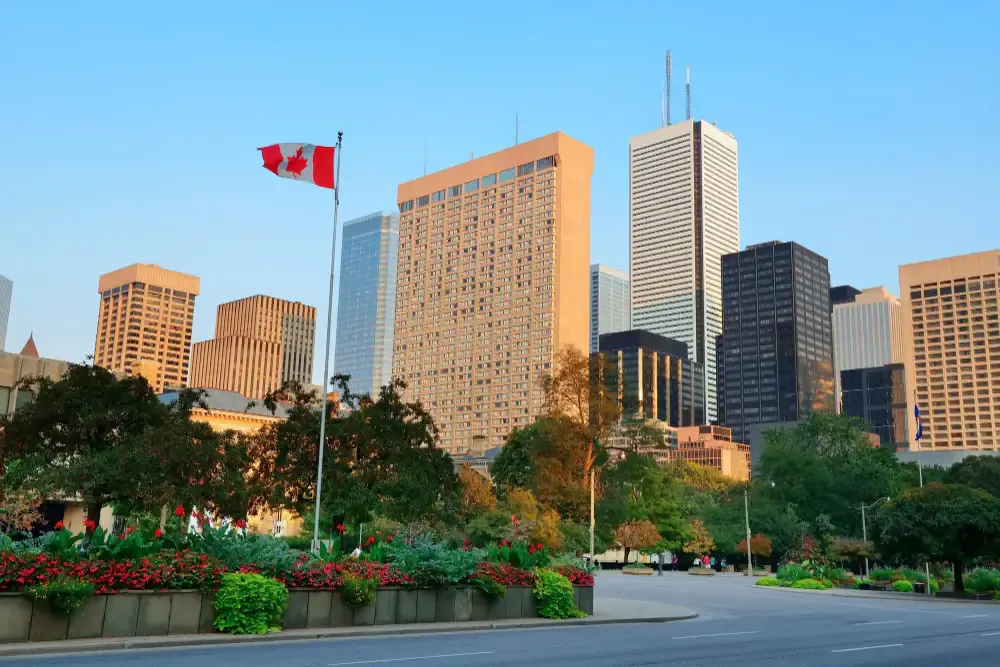 Vista de arranha-céus em uma avenida, com bandeira do Canadá ao alto e paisagismo com árvores e canteiros floridos em primeiro plano sob céu azul.