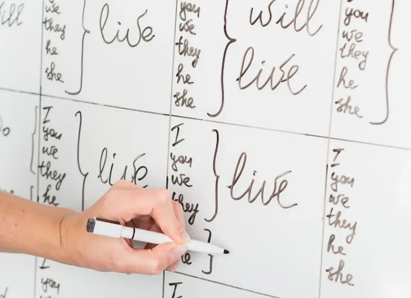 Pessoa escrevendo em quadro branco com marcador e caneta, formando frases em inglês como “you”, “we” e “they he she live”, com mão segurando a escrita