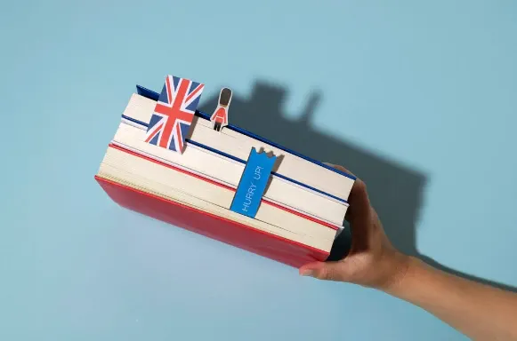 Mão segurando uma pilha de livros com capa vermelha e bandeira do Reino Unido, com marcador vertical escrito “Hurry up” sobre fundo azul claro e sombra ao lado.