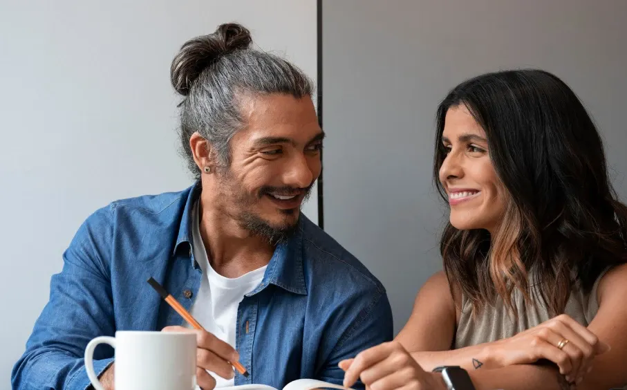 Casal sorridente tomando café em casa: homem com cabelo grisalho e barba usando camisa jeans e mulher de cabelo longo conversando e gesticulando sobre a mesa, com xícaras e copo ao fundo.