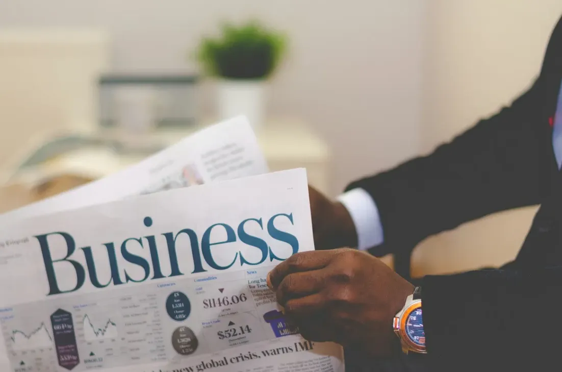 Pessoa sentada em um ambiente de trabalho lendo jornais financeiros com a manchete “Business”. Close mostra mãos segurando as folhas e gráfico ao fundo, sugerindo notícias do mercado e economia.
