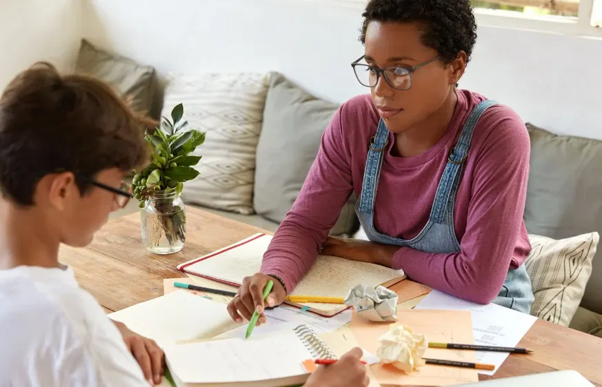 Duas pessoas estudando juntas em uma mesa de madeira, com cadernos e lápis coloridos, uma delas usando óculos e avental jeans, em ambiente interno com sofá ao fundo.