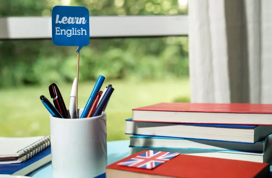 Pilha de livros didáticos e canetas em um ambiente ao ar livre, com um suporte azul escrito “Learn English” ao lado de material de estudo.