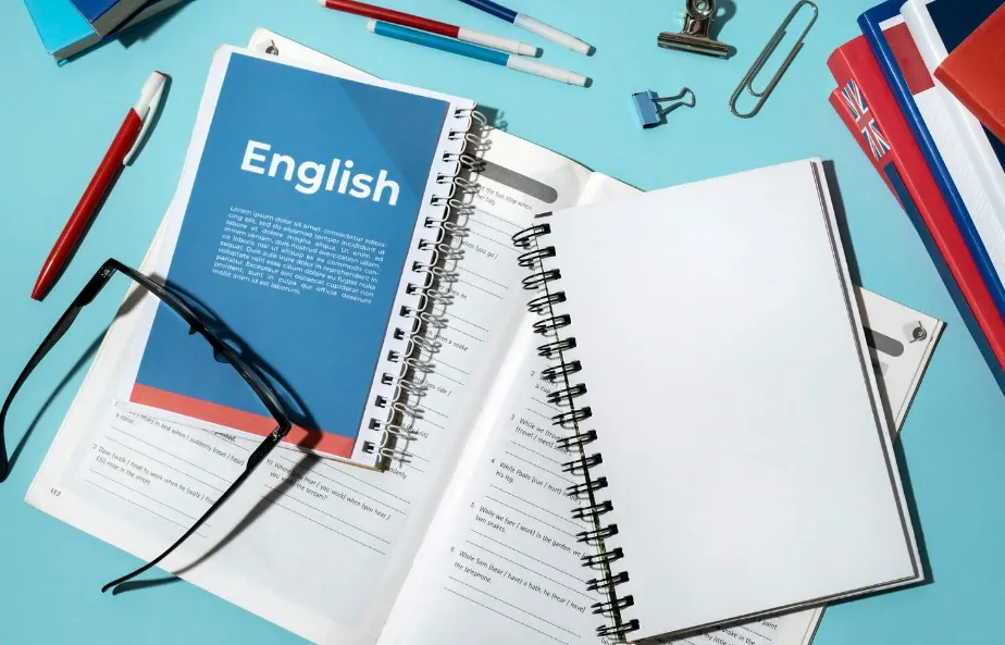 Materiais de estudo para aprendizado de inglês sobre uma mesa azul: livros abertos com texto “English”, caderno com espiral e lápis e canetas ao redor.