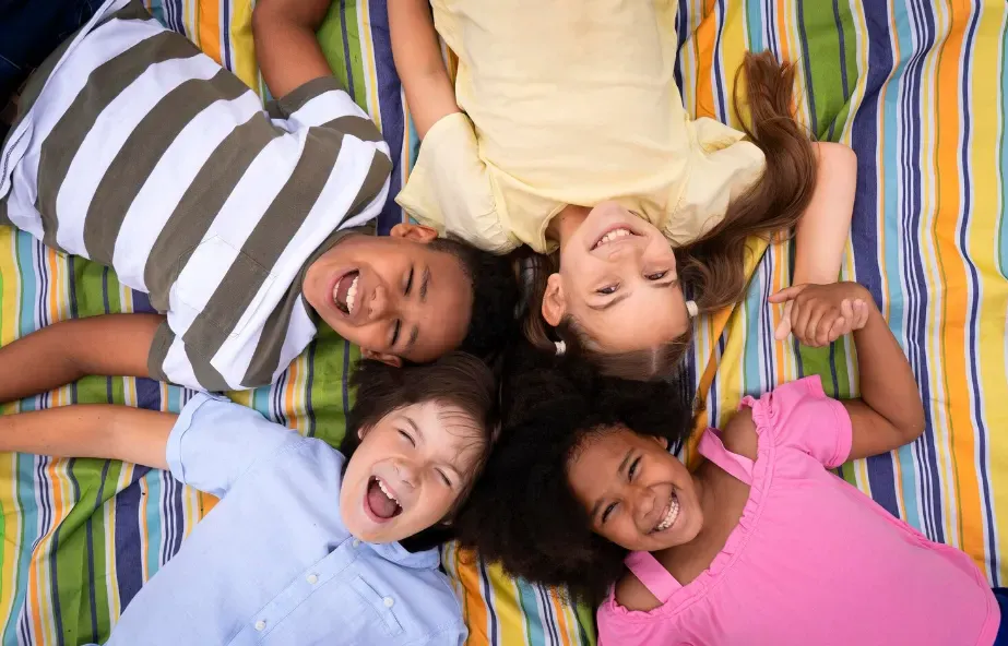 Foto de quatro amigos deitados lado a lado em um lençol estampado colorido, com expressões felizes e rindo, vista de cima compartilhando um momento descontraído.