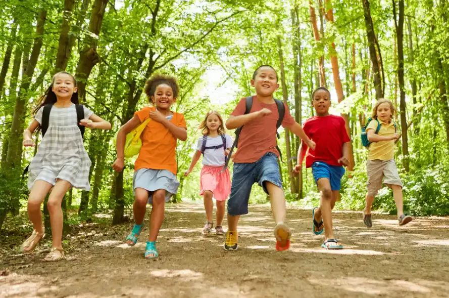 Grupo de crianças correndo e brincando em trilha de parque arborizado, com árvores verdes ao fundo e clima de diversão ao ar livre.