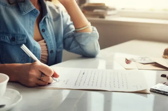 Pessoa escrevendo com caneta em uma folha de papel sobre uma mesa, em um ambiente com café e objetos de trabalho ao fundo, luz natural suave