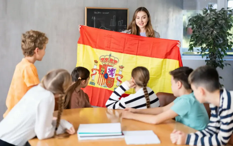 Crianças em sala de aula observam uma professora enquanto seguram e levantam a bandeira da Espanha, com o símbolo nacional ao centro.