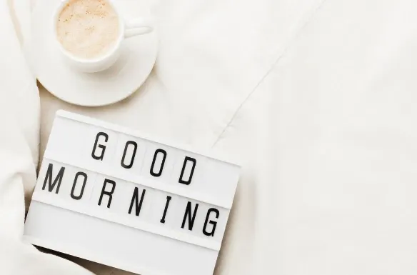 Café em xícara sobre pires e letreiro de marcenaria com a palavra Good Morning sobre tecido claro, em uma composição minimalista para mensagem matinal.