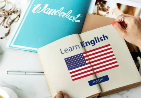 Livro aberto com a frase “Learn English” e bandeira dos Estados Unidos, sendo segurado por mãos sobre uma mesa; ao fundo, página azul com texto “Wonderful”.