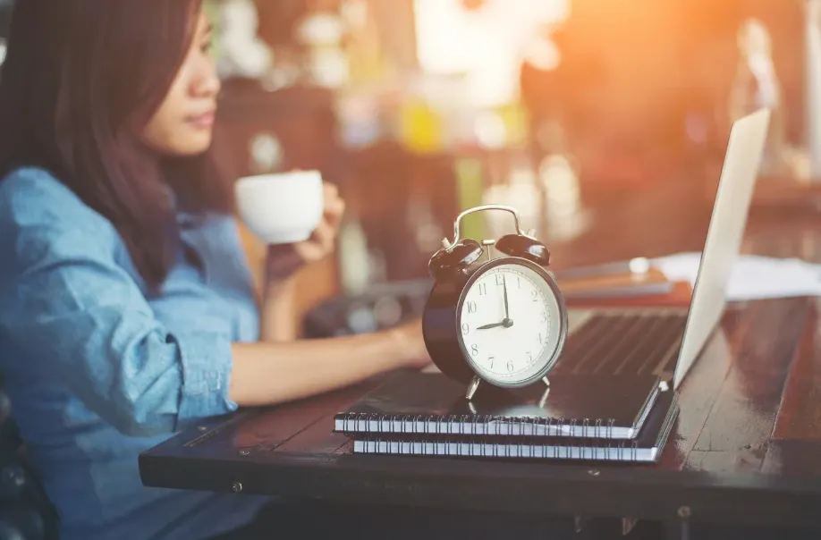 Mulher trabalhando em casa com notebook, enquanto um despertador marrom em primeiro plano destaca foco no tempo e na produtividade durante o trabalho.