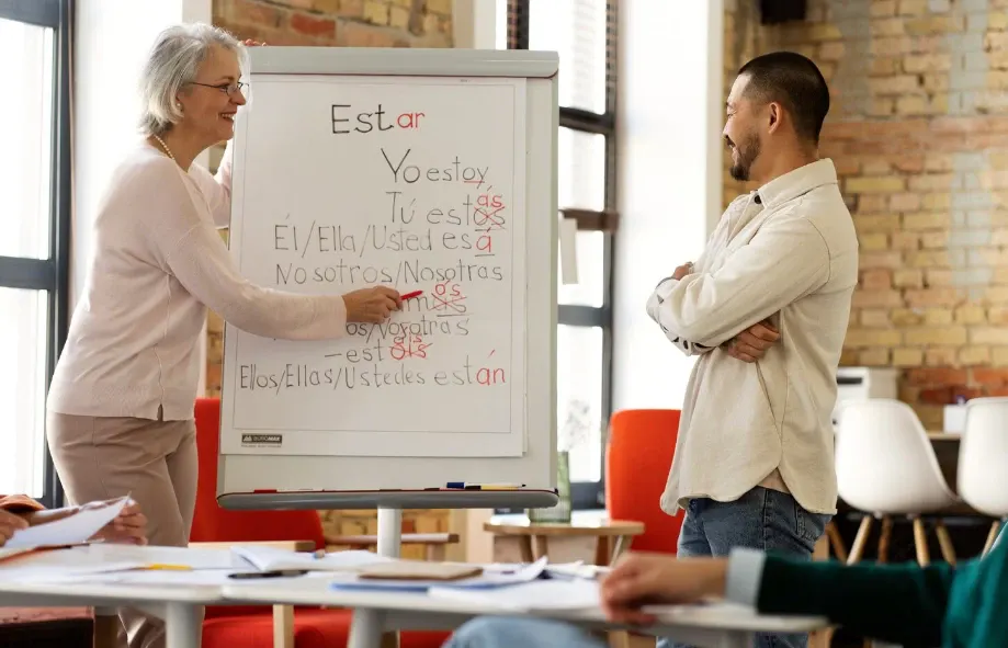 Professora aponta para um quadro branco com anotações em espanhol sobre pronomes e posições gramaticais, enquanto alunos discutem em sala de aula com parede de tijolos e mesas.