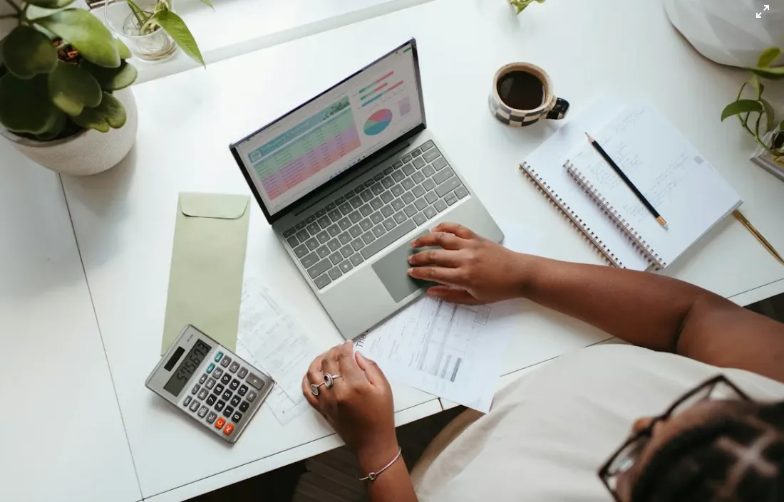 Mesa de escritório com laptop exibindo gráficos, calculadora, papéis e caneta, enquanto uma pessoa analisa documentos e faz anotações com café ao lado.