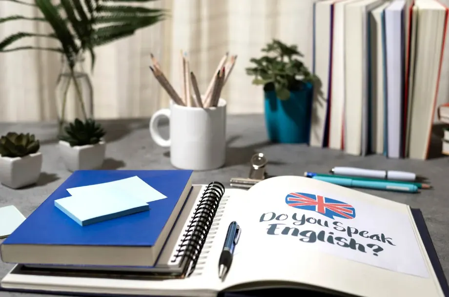 Mesa de estudos com caderno aberto e caneta, livros ao fundo e vaso com plantas; aparece o texto “Do You Speak English?” na página com bandeira.