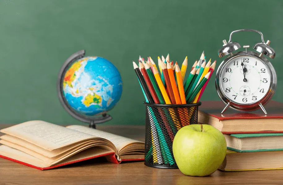 Escola e estudo: globo terrestre, lápis coloridos em um suporte metálico, maçã amarela, relógio de despertador e livros abertos sobre mesa com quadro verde ao fundo.