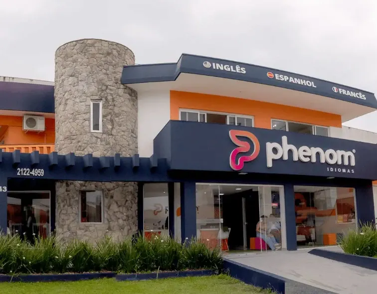 Fachada de loja com placa de opções de idiomas e logotipo phenom IDIOMAS, em prédio comercial com entrada e jardim frontal.