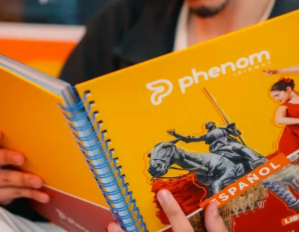 Capa amarela de livro escolar “phenom” com ilustração de soldados e título “SPANIOL”, enquanto mãos seguram um caderno com espiral azul ao lado, em ambiente de estudo.