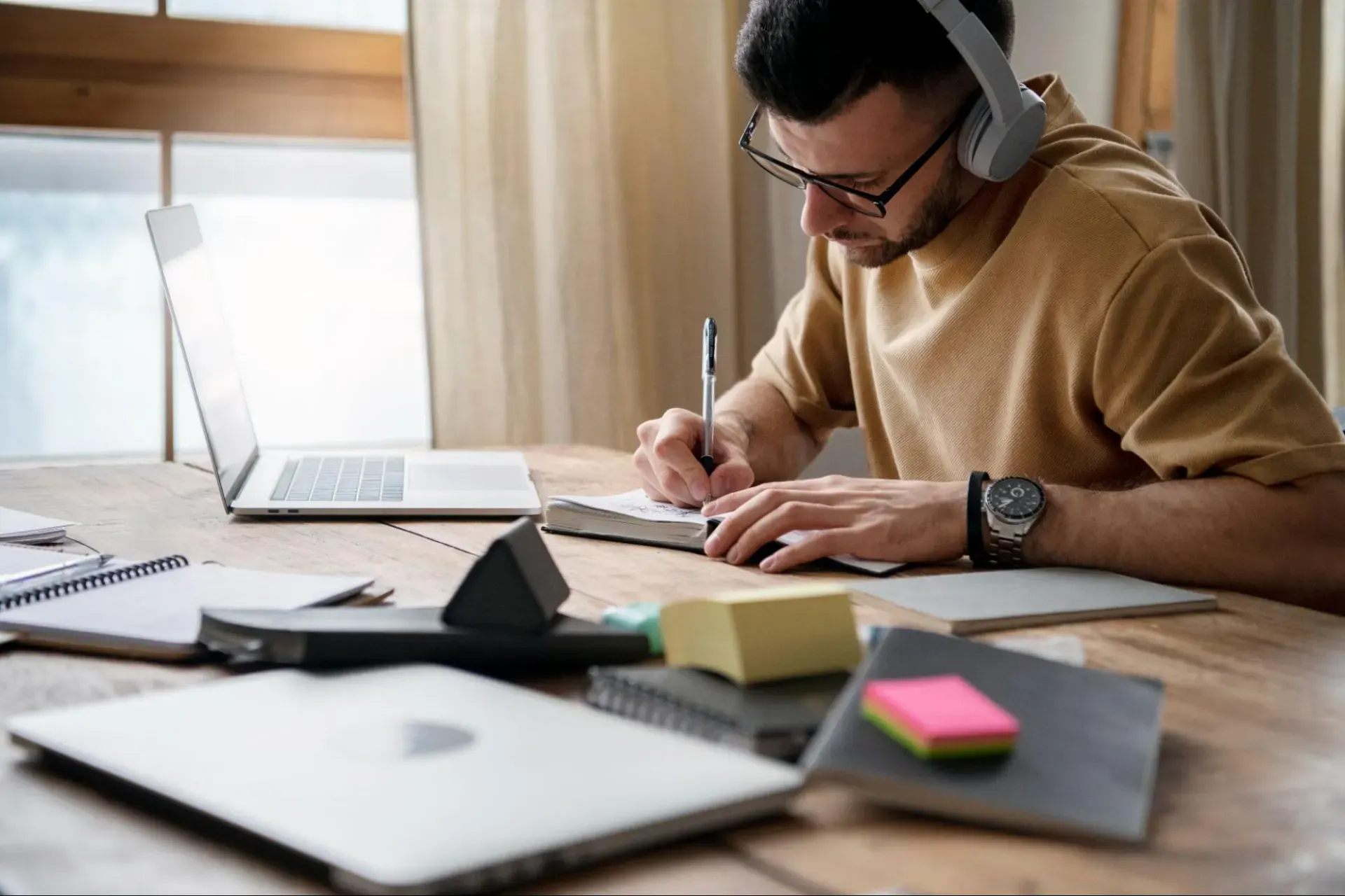 Homem trabalhando em home office com notebook, usando fones de ouvido e escrevendo em um caderno sobre a mesa, com materiais de estudo e celular ao lado.