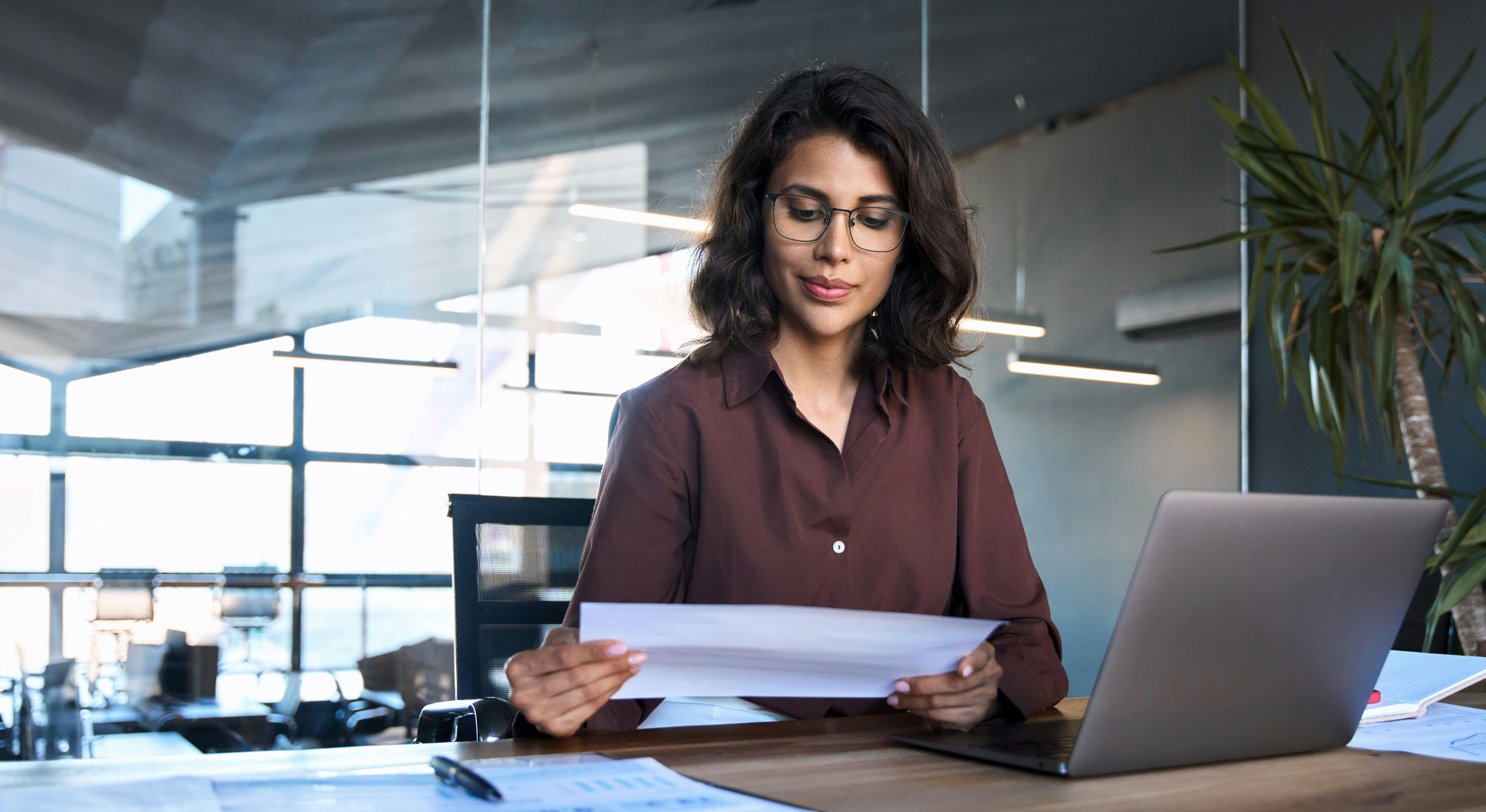 Mulher sentada, trabalhando em um escritório, analisando um papel, apoiada em uma mesa com o notebook aberto na sua frente.