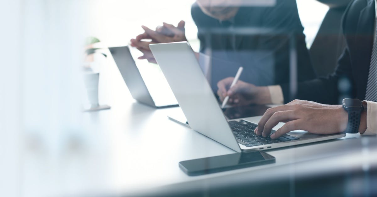 Homem digitando no trabalho, enquanto outro o observa apoiado em uma mesa com seus notebooks abertos na frente deles.