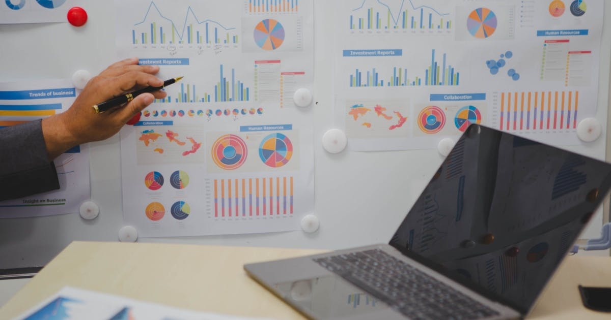 Homem mostrando gráficos na parede para ensinar a como organizar as finanças da empresa com o notebook aberto em cima da mesa