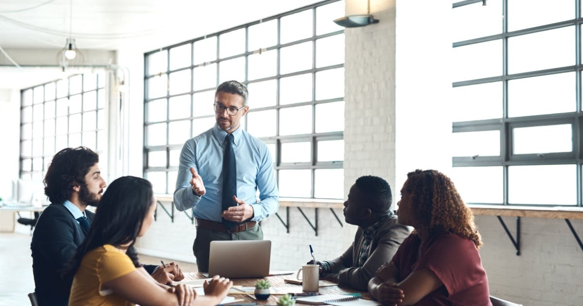 Homem de pé exercendo liderança e gestão com a sua equipe que está sentada em volta de uma mesa, enquanto ele fala.