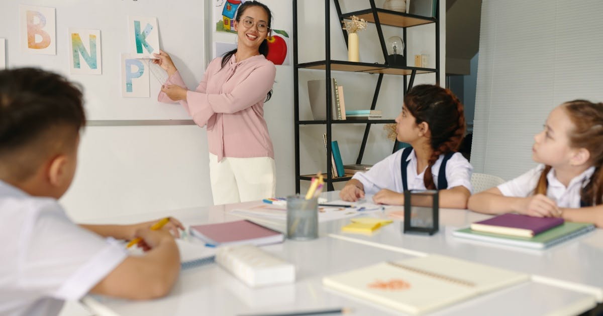 Professora em pé apontando para a lousa, dando aula em escola de inglês infantil, dando explicação para seus alunos sentados.