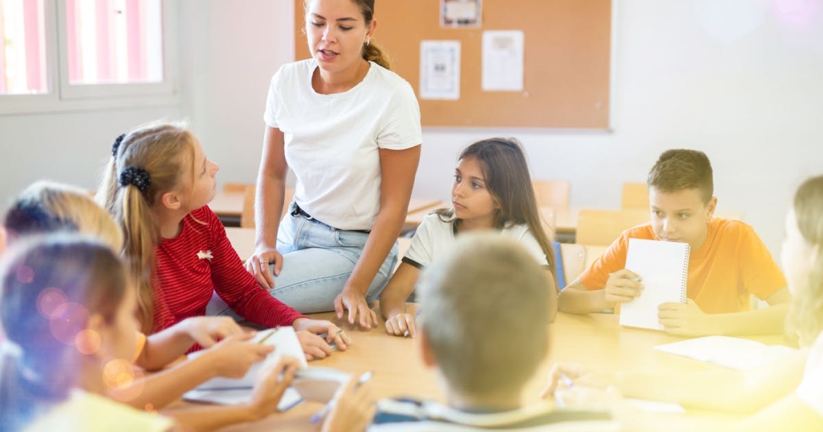 Professora sentada na mesa com seus alunos sentados em volta, dando aula em uma escola de inglês infantil.