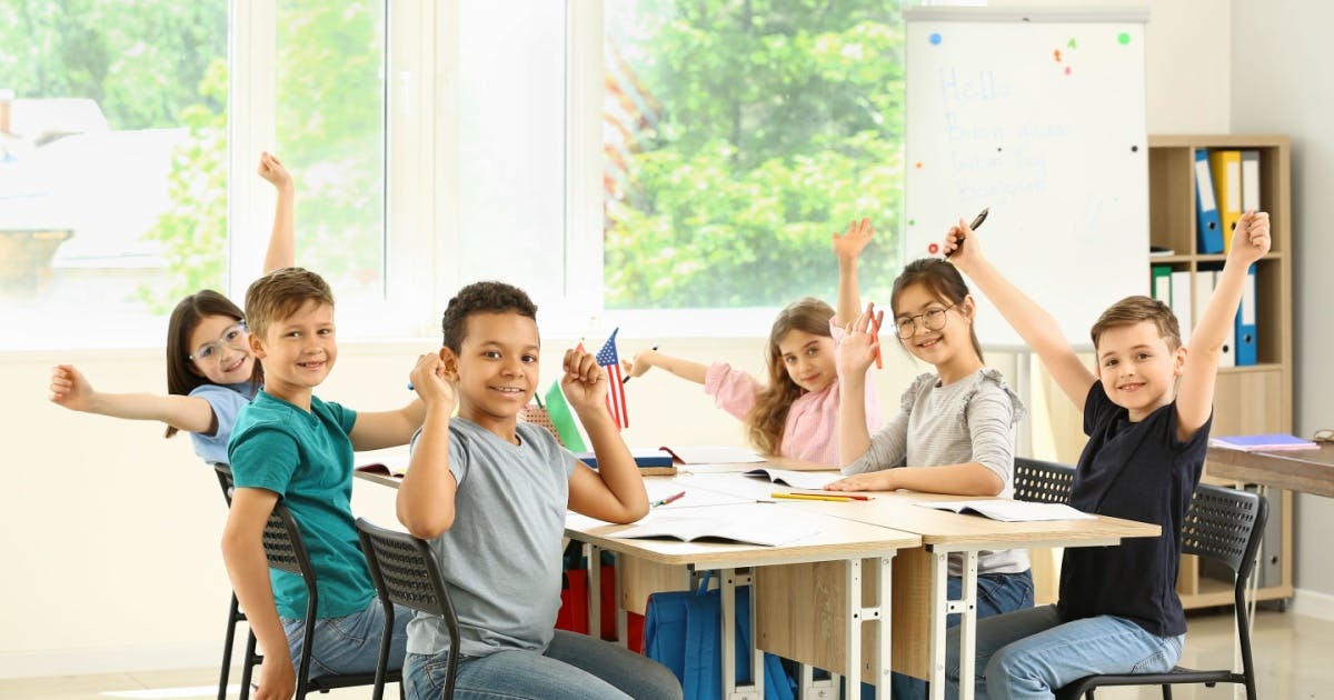 Alunos em uma sala de aula de uma escola de inglês infantil sentados, com papéis em cima das mesas, sorrindo enquanto vibram.