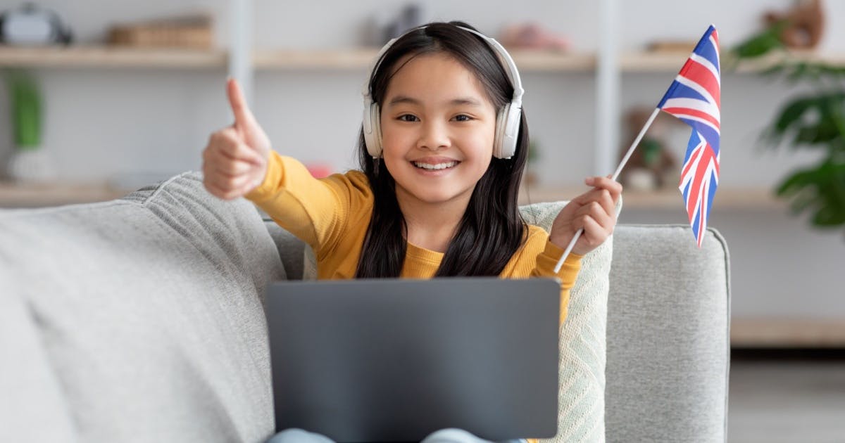 Menina sorrindo, com bandeirinha da Grã-Bretanha, usando fones e um notebook enquanto faz lição da escola de inglês infantil.