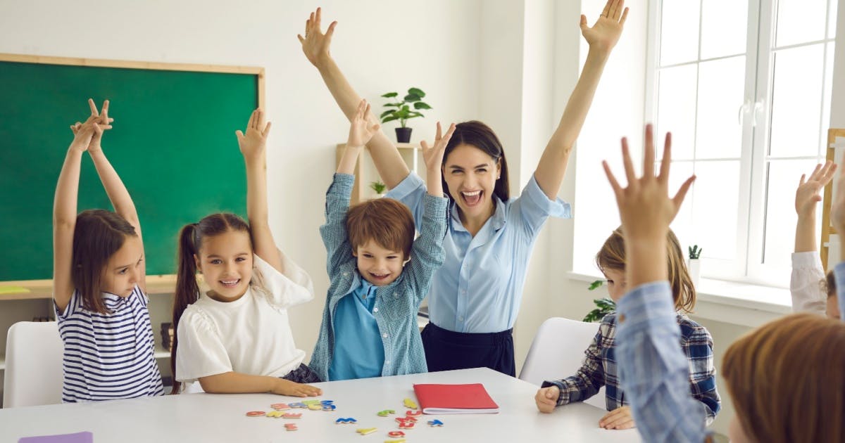 Crianças e professora vibrando em volta de uma mesa de uma sala de aula durante uma aula em uma escola de inglês infantil.