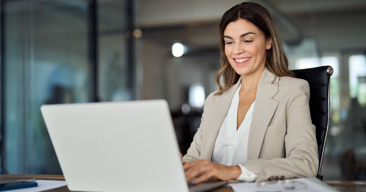 Mulher sentada e sorrindo, enquanto trabalha com microfranquias em um escritório, digitando em um notebook aberto.